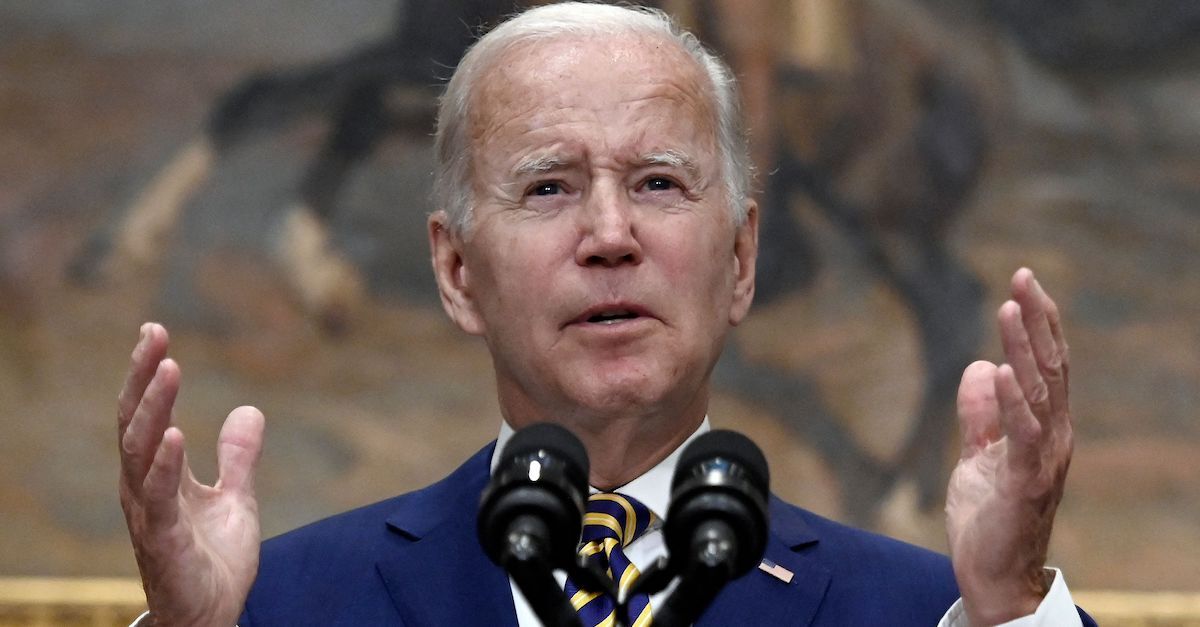 President Joe Biden is seen wearing a blue suit and white shirt, standing before microphones with his hands up as if to emphasize a point.