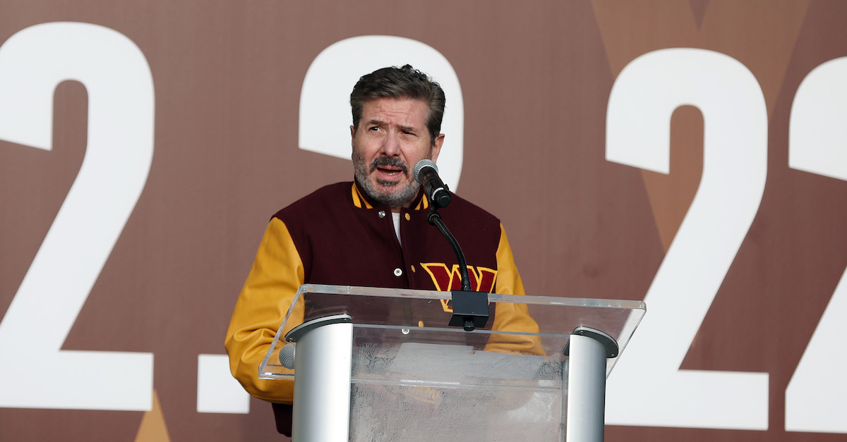 Washington Commanders co-owner Daniel Snyder is standing behind a silver podium with an attached microphone. He is wearing a varsity-style jacked with yellow sleeves and a dark red or maroon bust, with the letter "W" embroidered on the jacket's left side. He is speaking into the microphone.