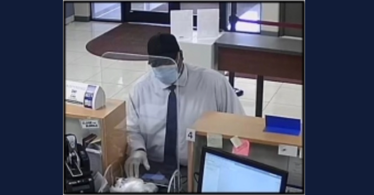 Nacoe Ray Brown is seen wearing a blue disposable face mask, rubber cloves, a baseball cap, a light collared shirt, and a dark tie. He appears to be communicating with a bank employee from behind a counter. 