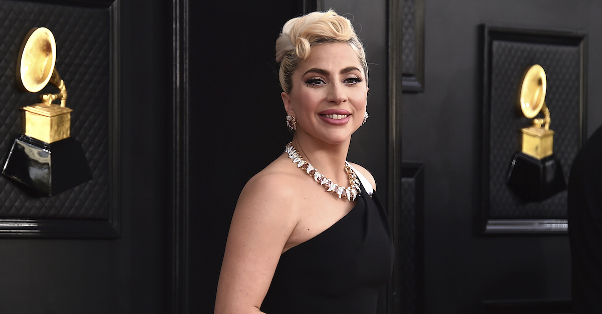 Lady Gaga wears a black one shoulder dress with a white stripe down the side. She stands in front of a wall with pictures of the "Grammy Awards" Picture.