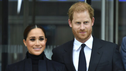 Meghan Markle is wearing a dark turtleneck and her hair is pulled back. She is standing next to Prince Harry, who is wearing a dark suit and tie with a white shirt.