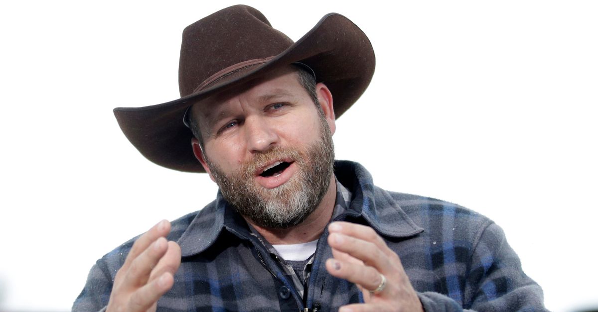 Ammon Bundy is seen wearing a blue and black flannel zip-up shirt and brown cowboy hat as he speaks. He appears to be gesticulating with his hands as he speaks.